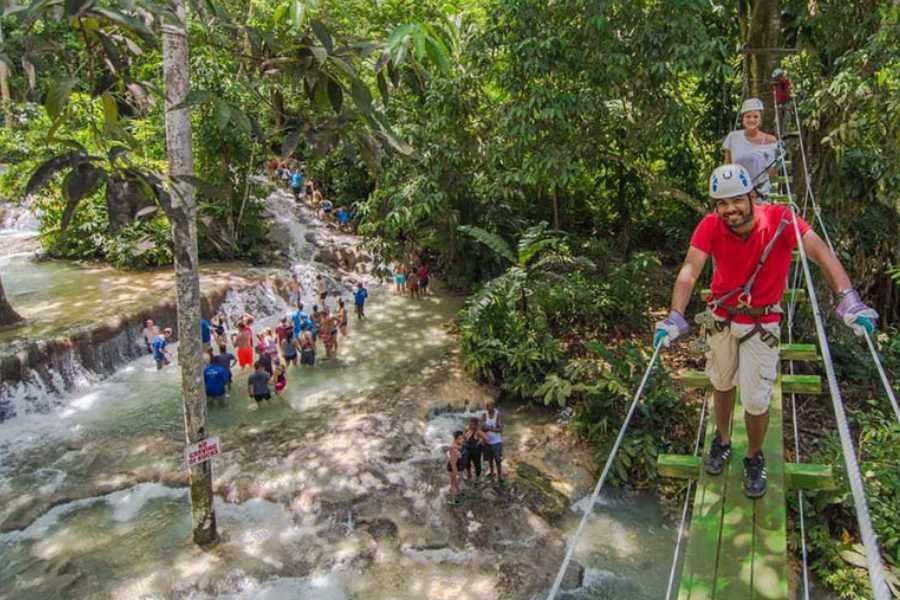 Adventurers cross a rope bridge above a waterfall in a lush, green forest.