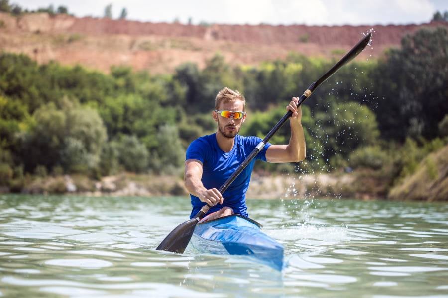 A young man is paddling a blue kayak on calm water.