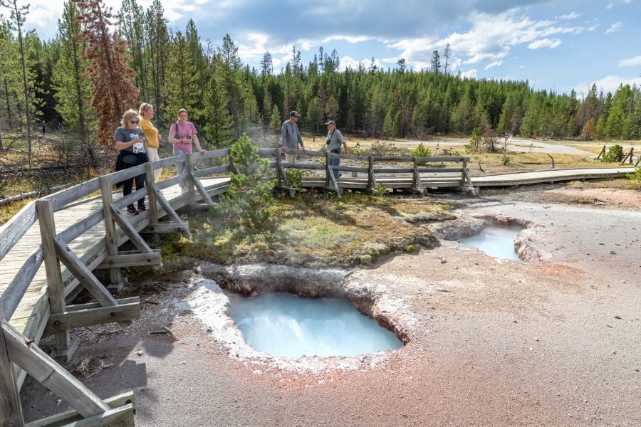 Visitors walking along a wooden boardwalk past steaming hot springs.