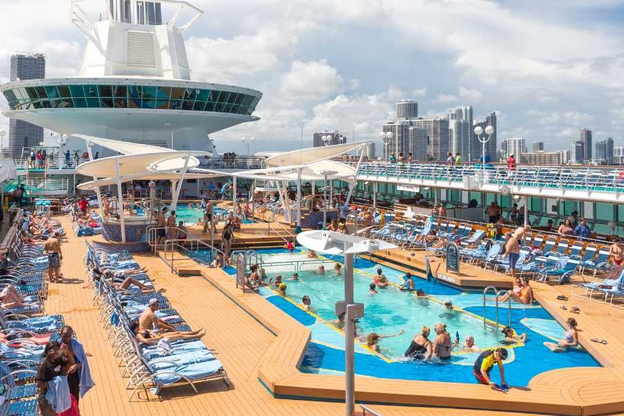 A lively cruise ship pool deck with sun loungers, swimming pools, and views of the nearby city skyline.