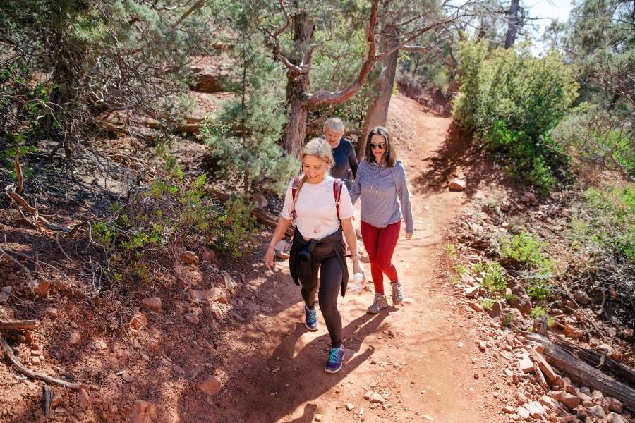 Three hikers walk uphill on a dirt path surrounded by red rocks and trees.