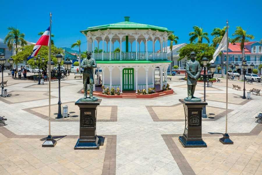 A picturesque town square with statues, a gazebo, and flags under the bright sun.