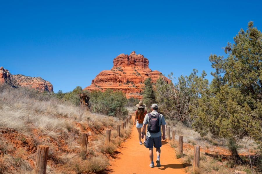 Hikers walk toward a famous red rock mountain on a sunny Sedona trail.