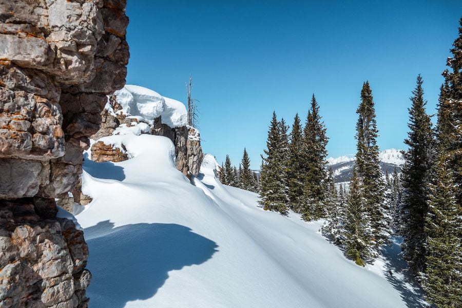 Fresh snow blankets rocky cliffs and tall evergreens on a clear winter day.