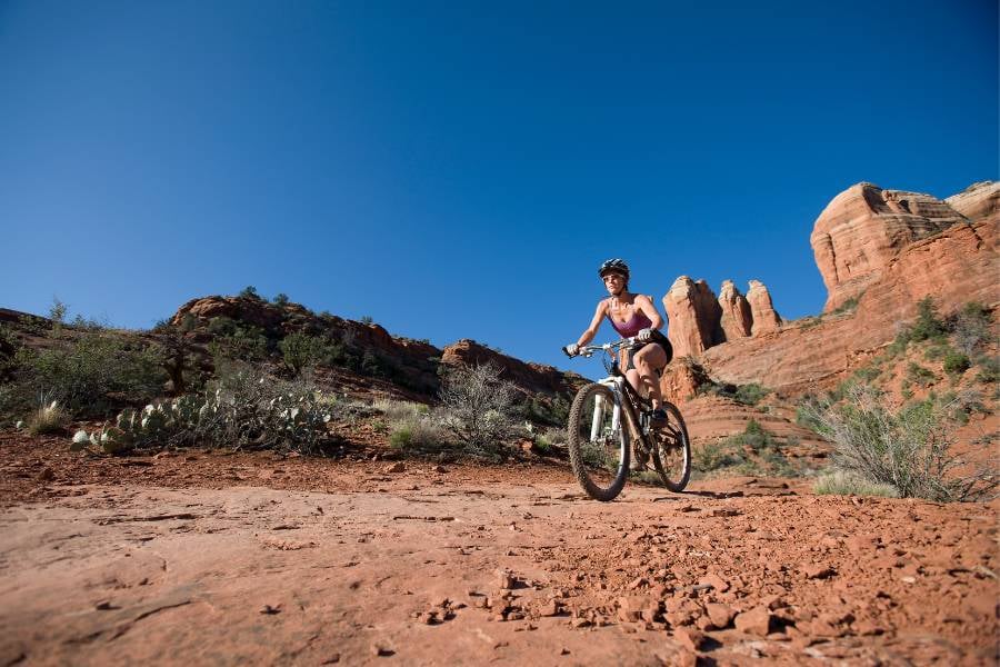A woman is riding a bike on a rocky red trail with blue sky and cliffs behind.