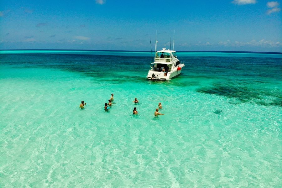 Travelers swimming in shallow, crystal-clear water near a small boat.