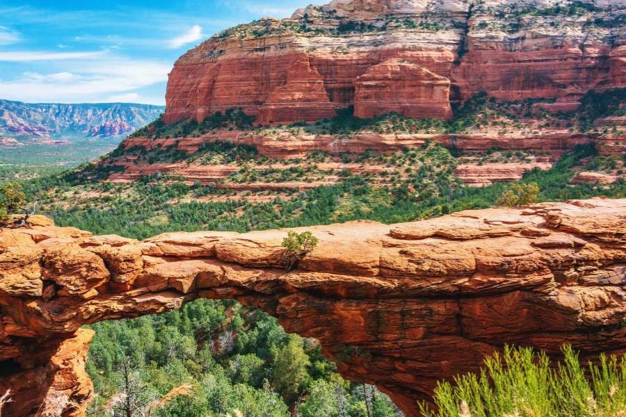 A natural red rock bridge stands high above a forested canyon.