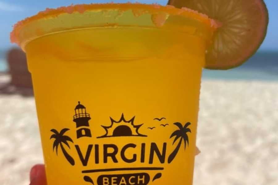 A bright orange tropical drink with a lime garnish, held against a backdrop of a sunny beach and ocean.