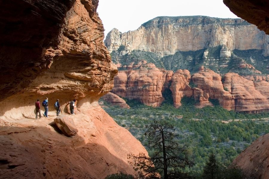 A group hikes along a ledge under red cliffs, with views of Sedona below.