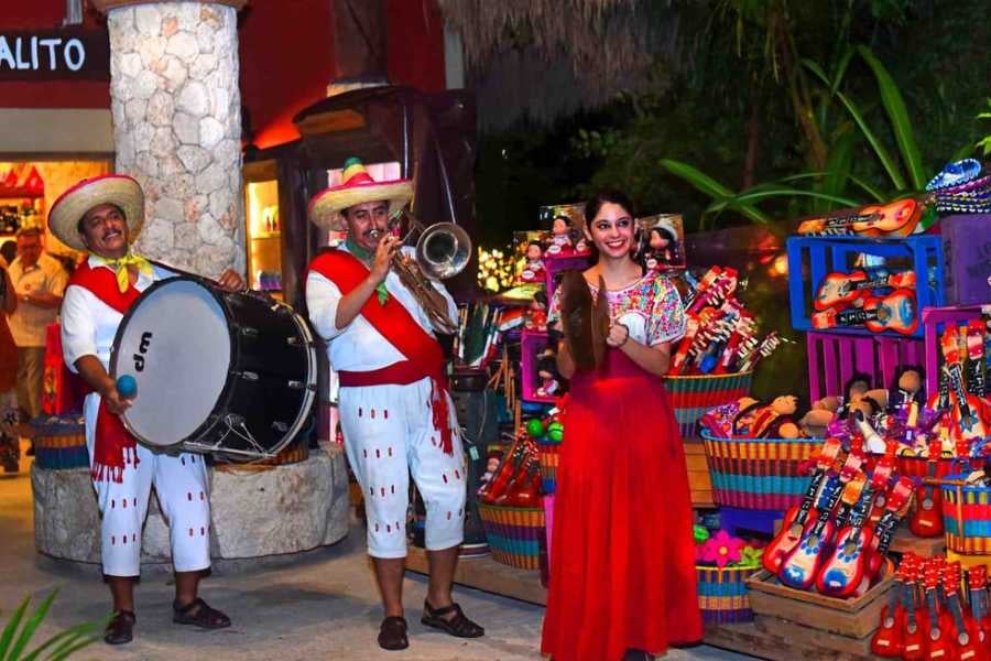 A cheerful band plays instruments as a woman in a red dress poses near colorful market stalls.