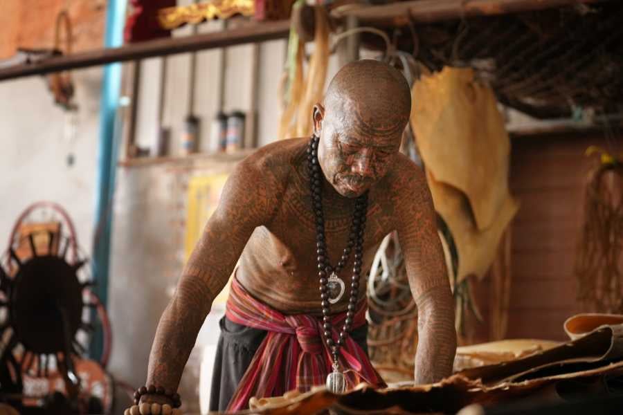 A tattooed artisan crafting traditional items, surrounded by cultural tools in a workshop setting.