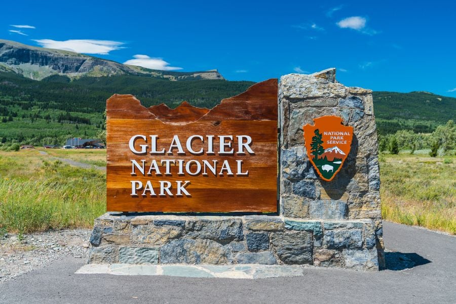 A wooden sign welcomes visitors to Glacier National Park under a bright blue sky.