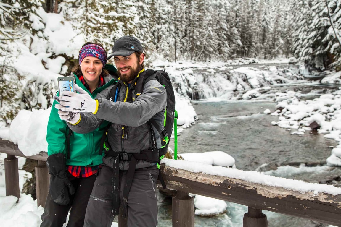 young couple taking picture on bridge with snowy river behind