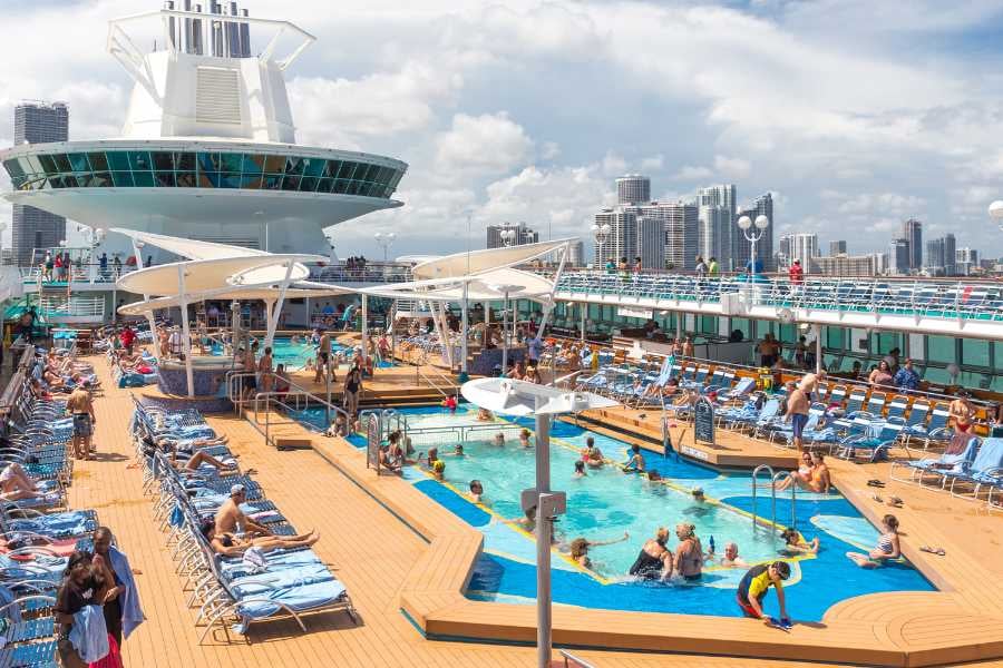 A busy pool deck on a cruise ship with people relaxing and city views behind.