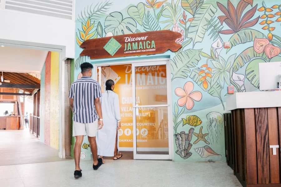 A couple walks into a vibrant "Discover Jamaica" center, decorated with tropical murals, ready for a new adventure.