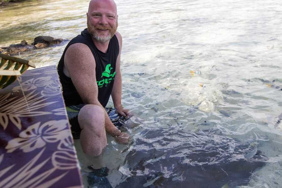 A smiling man kneels in shallow water, surrounded by tiny fish, enjoying a unique marine experience.