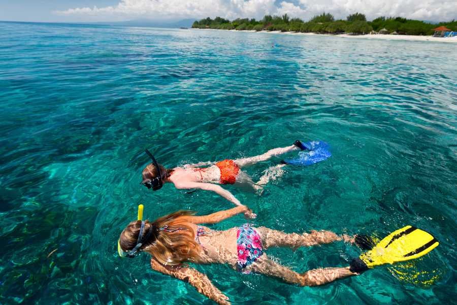 Two snorkelers explore a vibrant underwater world in stunningly clear blue water near a tropical shoreline.