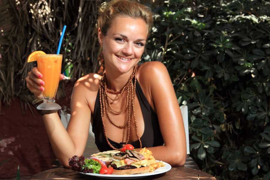  A happy woman sips a bright orange smoothie, enjoying a colorful meal in a lush outdoor setting.