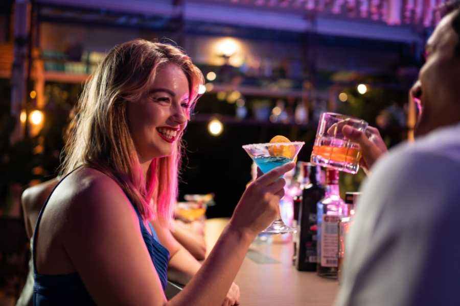 A woman smiles brightly while holding a cocktail, celebrating with a friend at a lively bar.