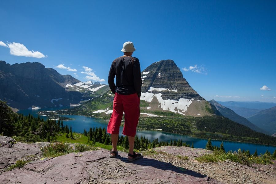 Hiker overlooking a lake surrounded by snow-capped peaks and green forests.