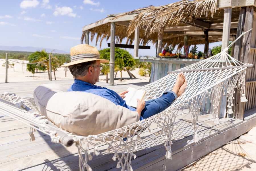 Relaxation perfected: A man in a straw hat reads in a cozy hammock under a beachside thatched roof.